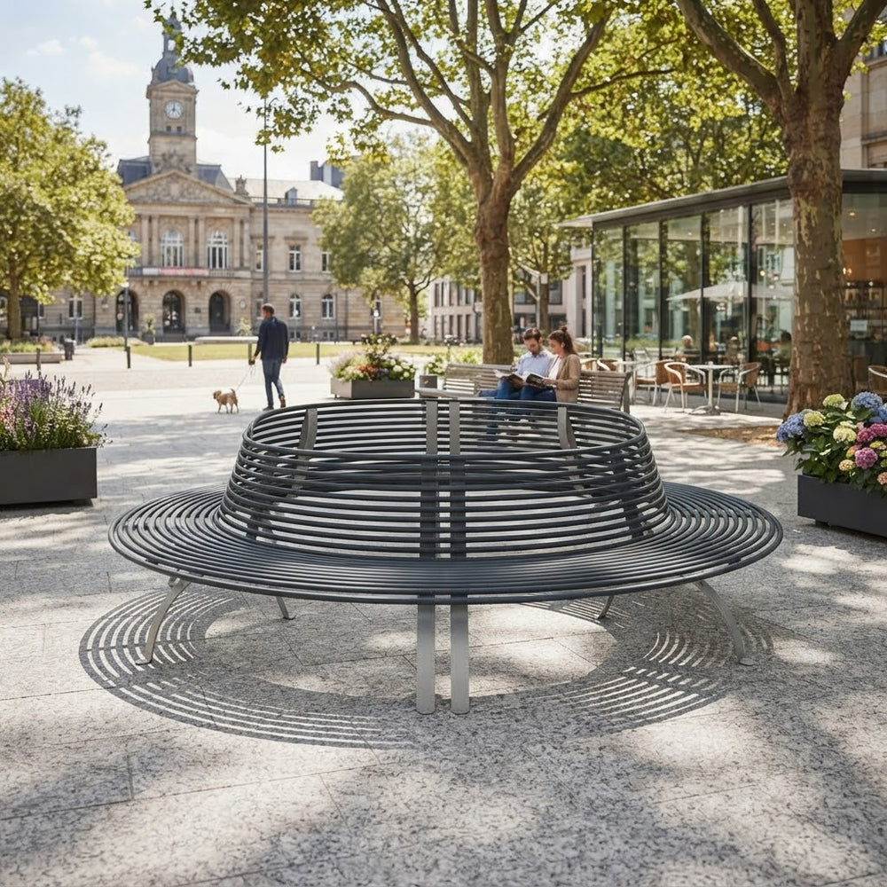 A modern circular metal park bench with a slatted back and seat, situated in a sunny urban plaza with trees, people, and a historic building in the background.