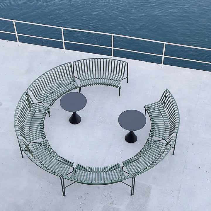 A curved outdoor seating set on a concrete deck overlooking the ocean, featuring a semi-circular metal bench with green slats, two round black tables, and a white railing above.