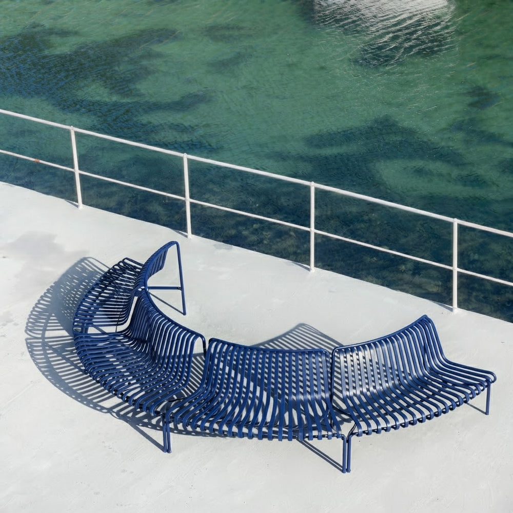 A curved outdoor metal bench in dark blue sits on a light concrete deck beside a railing overlooking greenish water.