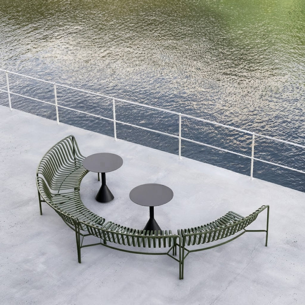 Outdoor curved metal patio seating set with two small round tables, placed on a concrete deck beside a body of water with a white railing.