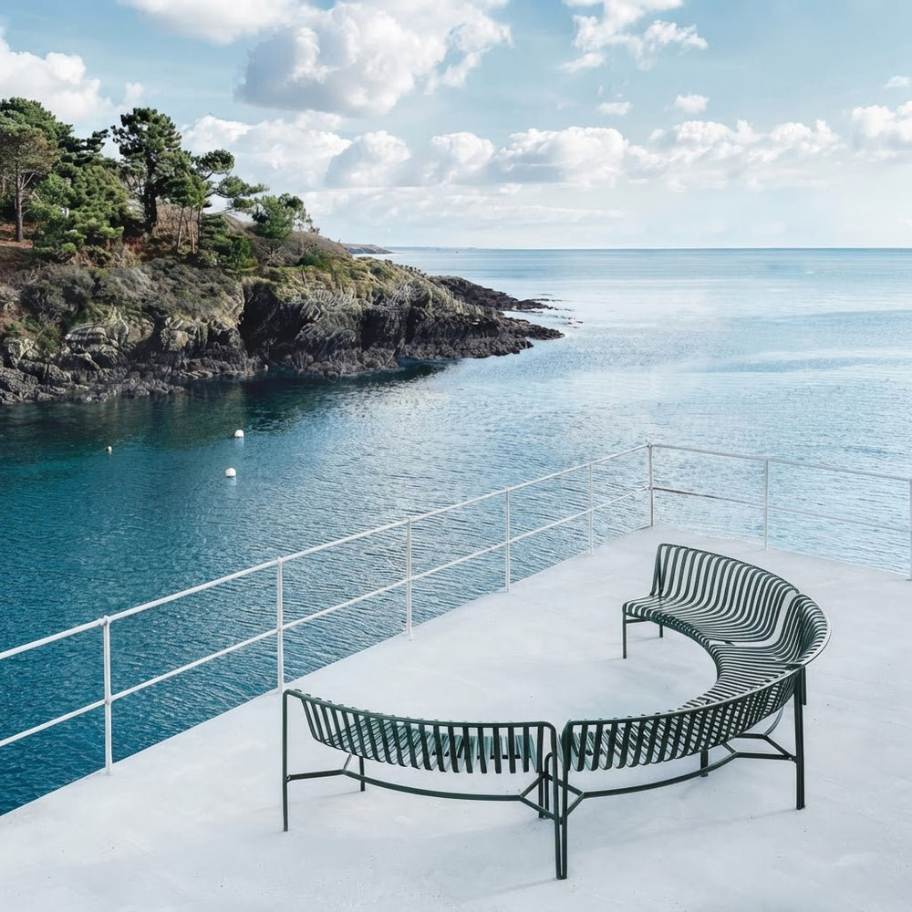 Two curved outdoor metal benches on a white concrete terrace overlooking a rocky coastline and blue sea under a cloudy sky.