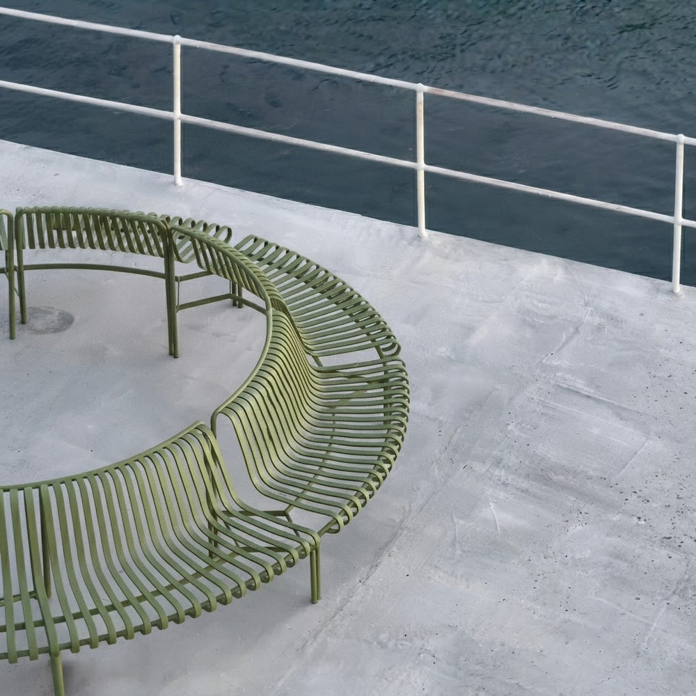 An olive green curved metal outdoor bench with a slatted design, situated on a concrete deck next to a white railing and dark water.