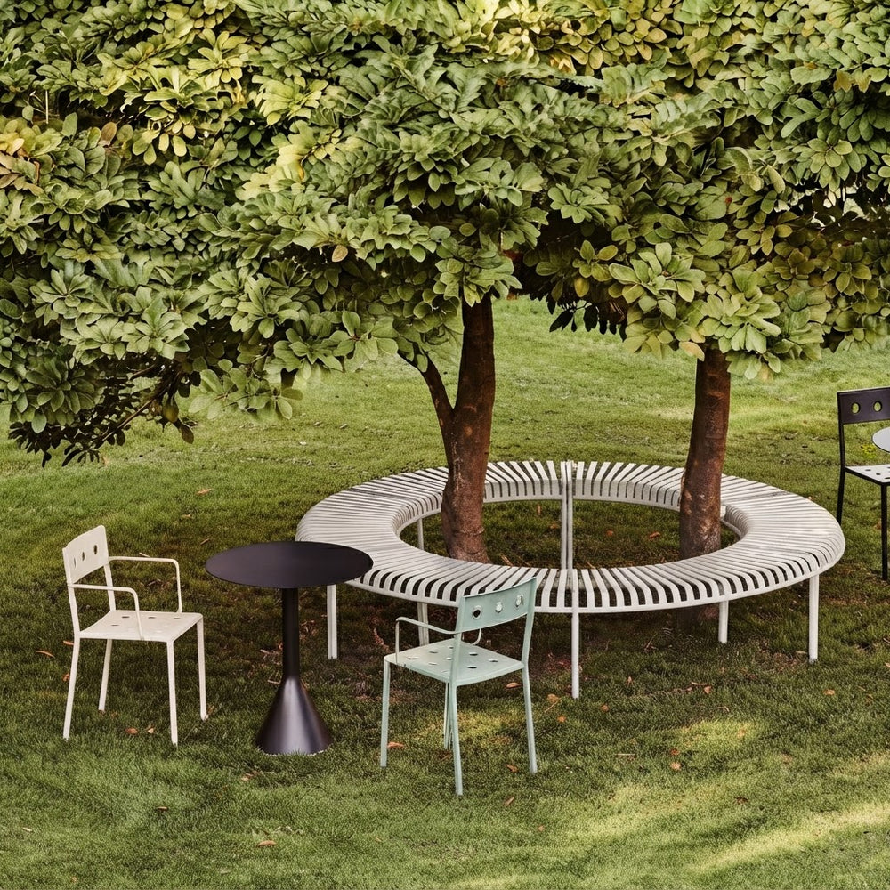 Outdoor patio furniture arrangement on a grassy lawn beneath a large leafy tree, featuring a curved white slatted bench, a small black round table, and two chairs in off-white and pale mint green.