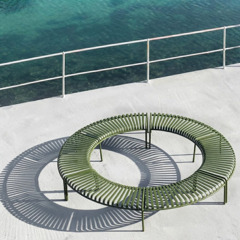 An outdoor circular metal lounge bench in olive green sits on a light concrete deck beside a railing overlooking clear turquoise water. The bench has a slatted curved design with visible support legs, and its shadow is cast sharply on the deck.