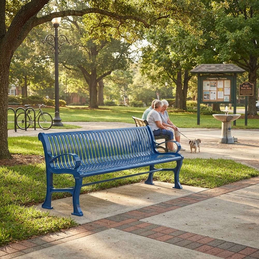A bright blue metal park bench in a tree-lined public park, with two people sitting on a bench in the background and a small dog nearby.