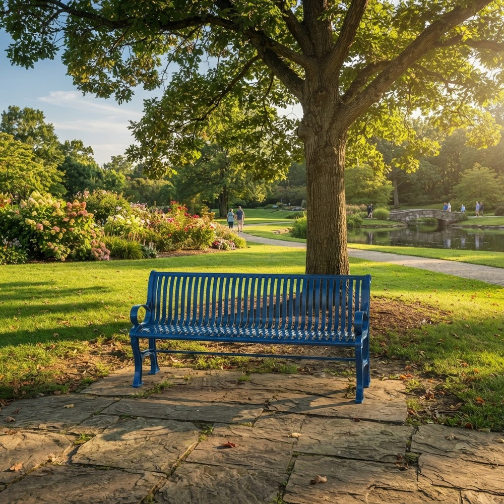 A bright blue metal park bench sits on a stone patio beneath a large tree in a sunny, green park with a pond and walking path in the background.
