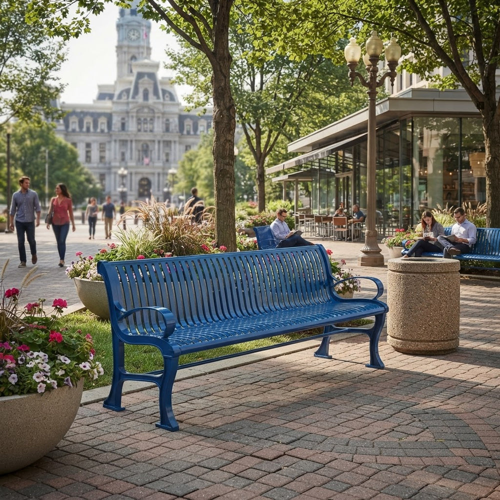 A bright blue metal park bench in a tree-lined urban plaza, with people sitting nearby and a large historic building in the background.