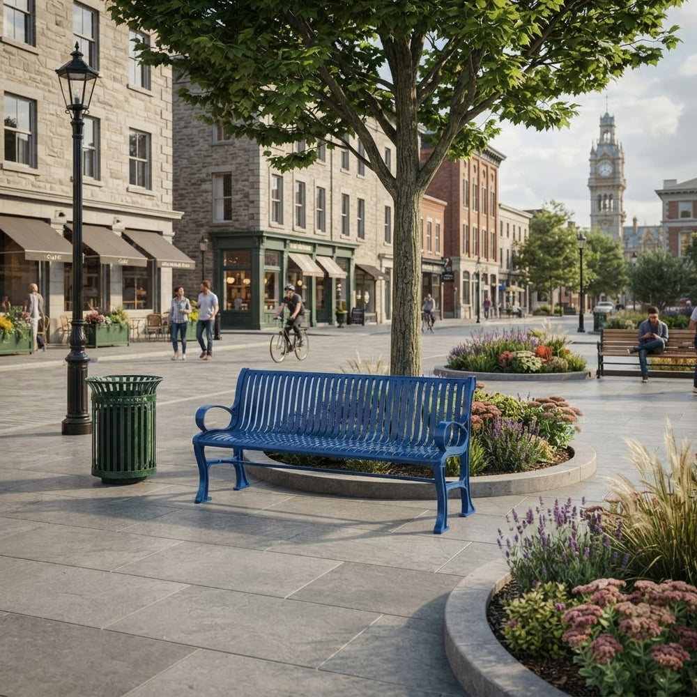 A bright blue metal park bench in a paved town square, surrounded by landscaped flower beds, a green trash can, a black lamppost, and a large tree. Historic stone and brick buildings line the street in the background, with a clock tower visible in the distance.
