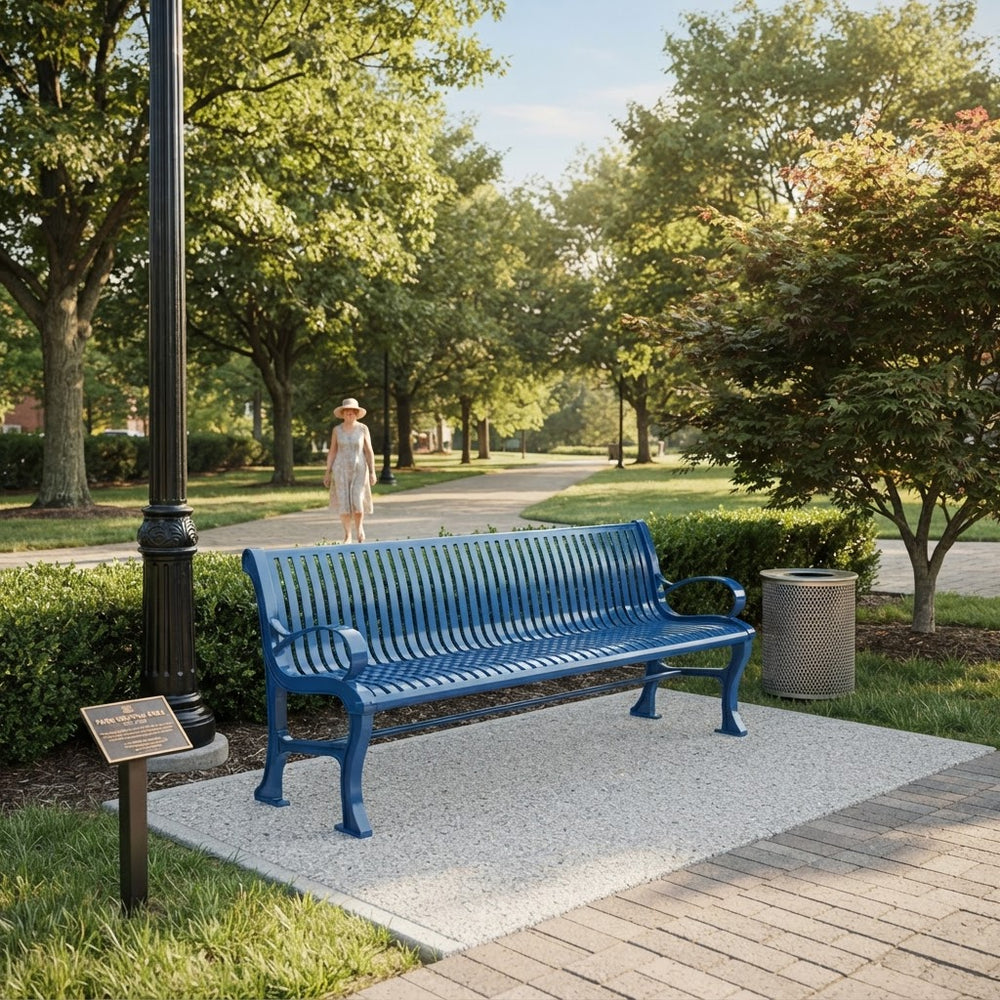 A bright blue metal park bench with slatted back and seat, placed on a light concrete pad beside a black lamppost in a landscaped park. A woman walks in the background along a tree-lined path, and a gray trash can sits to the right of the bench.