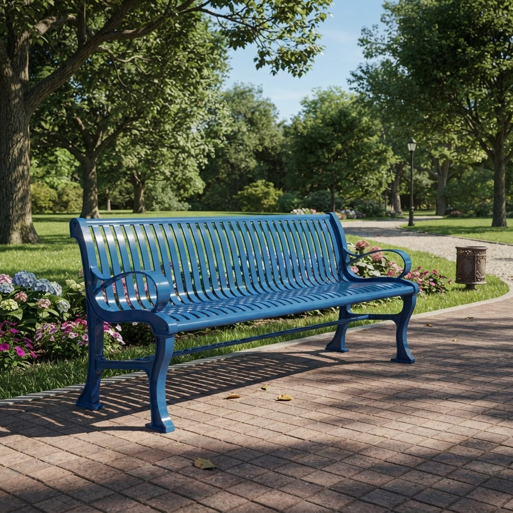 A bright blue metal park bench with curved armrests and a slatted back and seat, positioned on a paved pathway in a lush green park with trees and flowers.