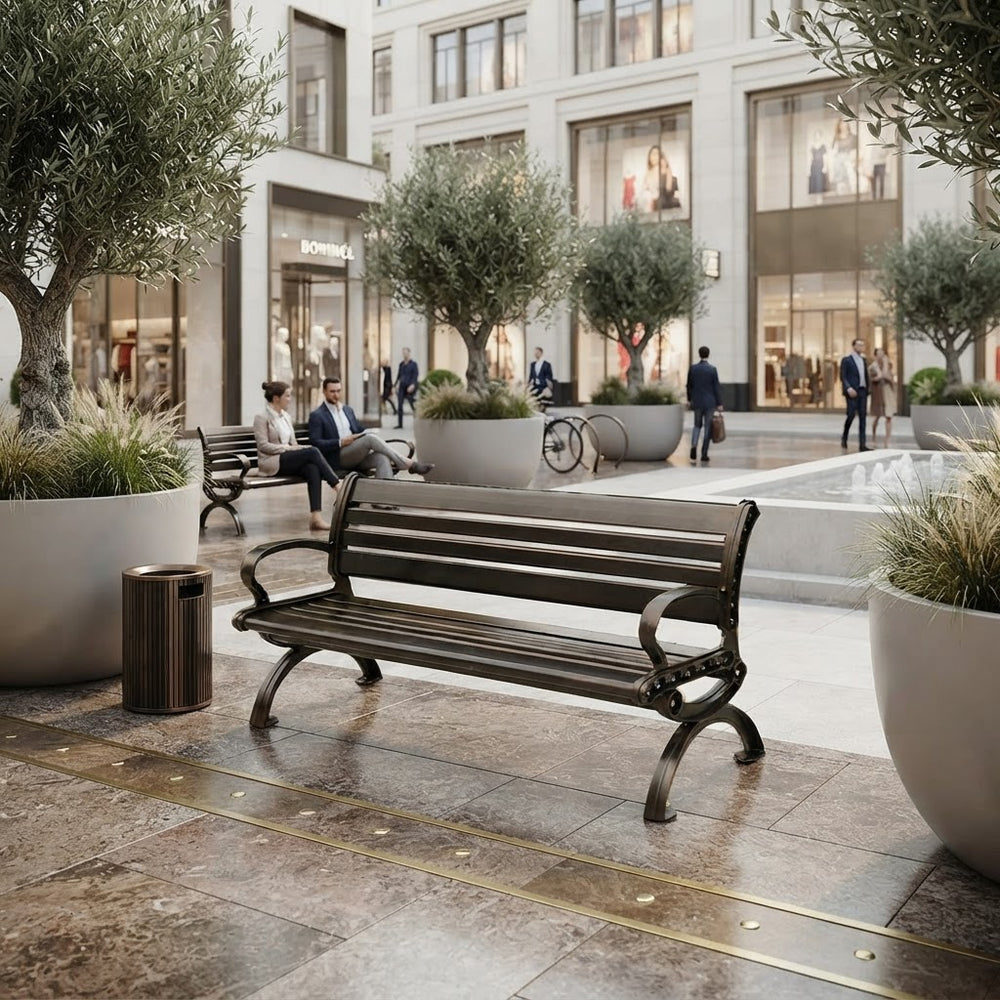 A dark brown wooden park bench with curved armrests and ornate metal legs, situated in a modern outdoor shopping plaza with large potted trees and stone tile flooring.