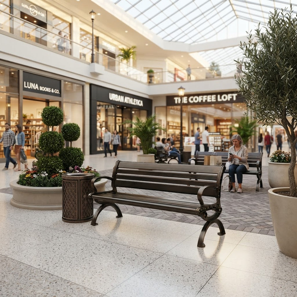 A dark brown wooden park bench with curved arms and legs stands in a modern shopping mall atrium with a glass roof, surrounded by potted trees, planters, and other shoppers.