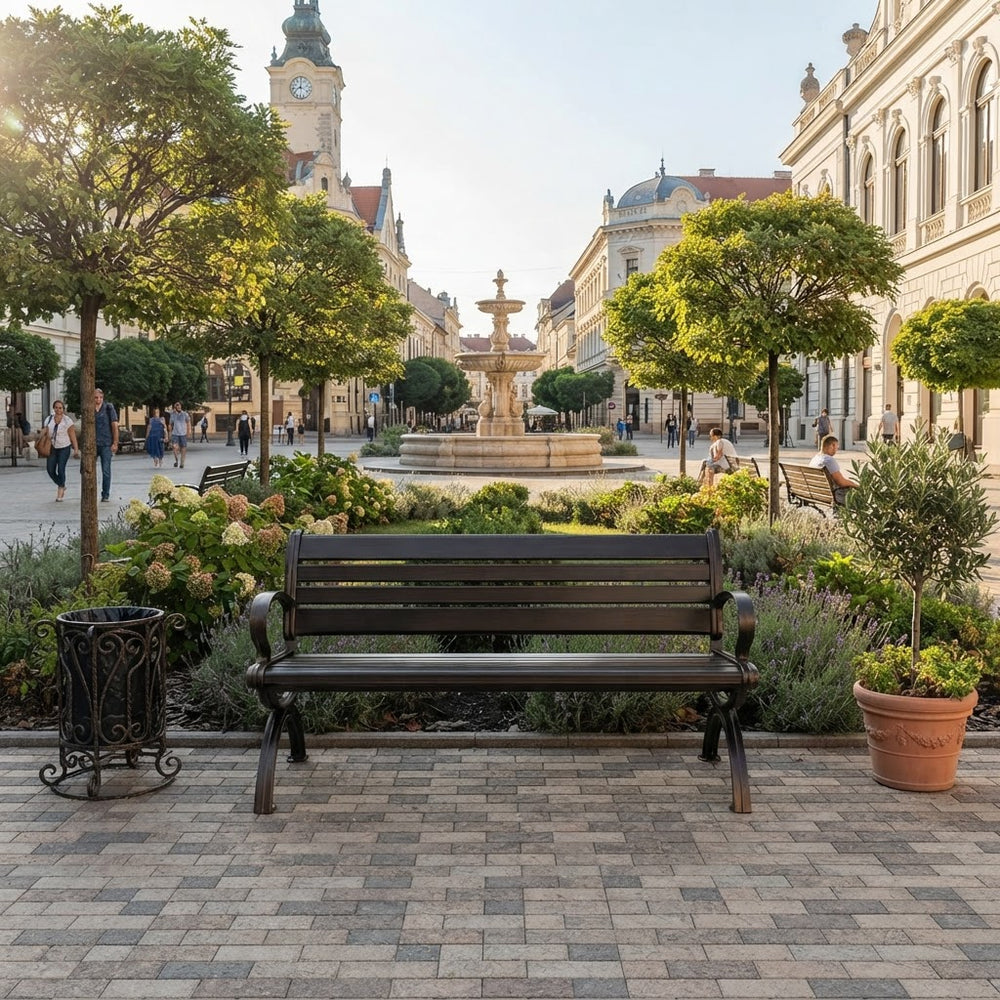 A dark brown wooden park bench with curved black metal arms sits on a paved plaza in a beautifully landscaped town square with a fountain, trees, and historic buildings in the background.