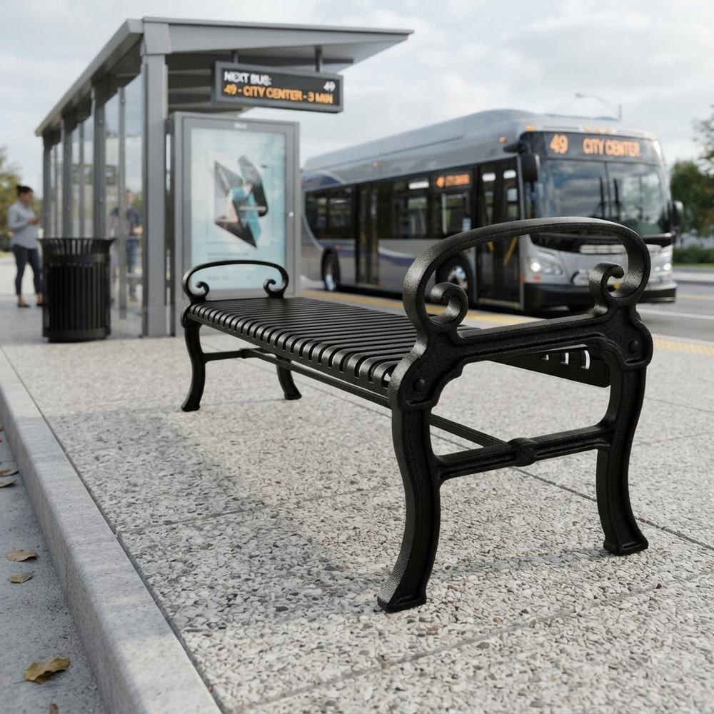 A black metal city bench sits on a concrete sidewalk at a bus stop, with a city bus and a shelter in the background.