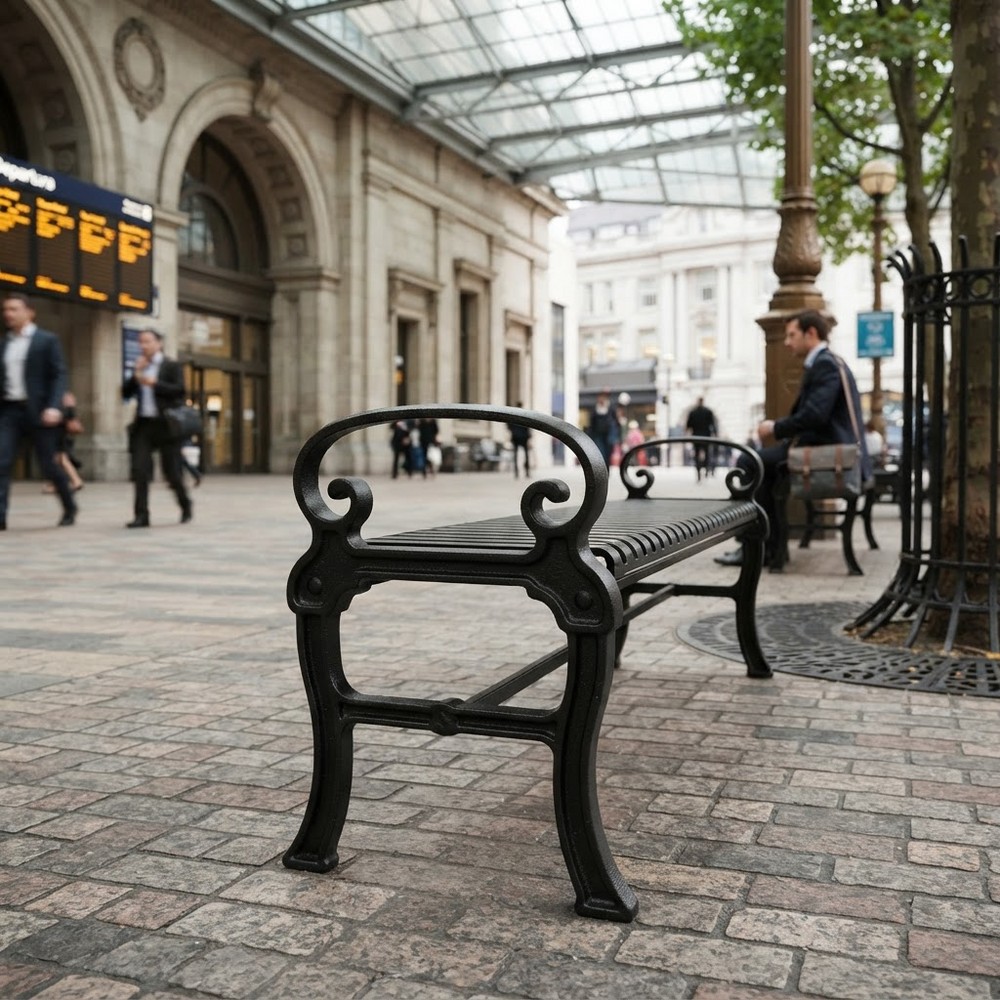 A black cast iron park bench with ornate scrollwork arms and legs, positioned on a stone-paved street under a glass-domed walkway with blurred pedestrians in the background.