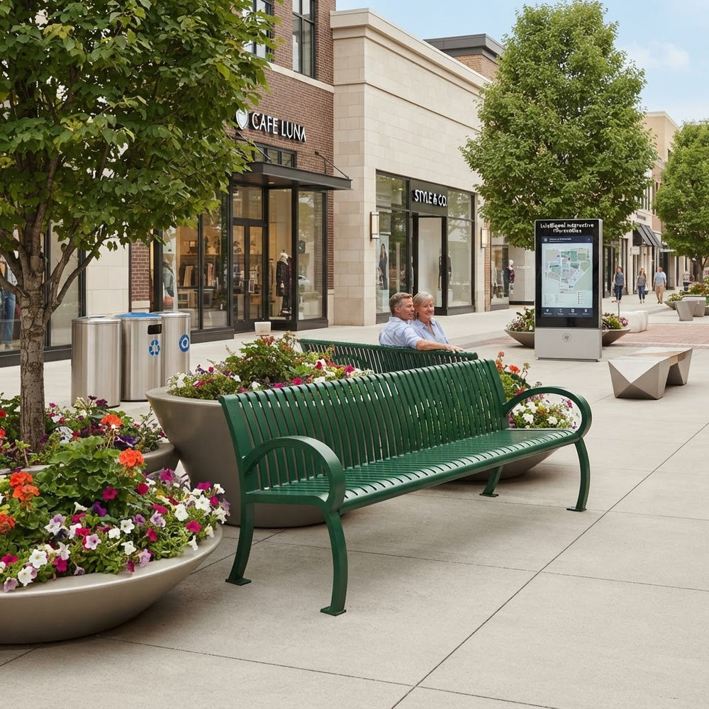A green metal park bench with curved armrests sits on a paved outdoor plaza in front of storefronts. Two people are seated on the bench, and large flower planters, trees, trash cans, and a digital map kiosk are visible in the surrounding shopping-area setting.