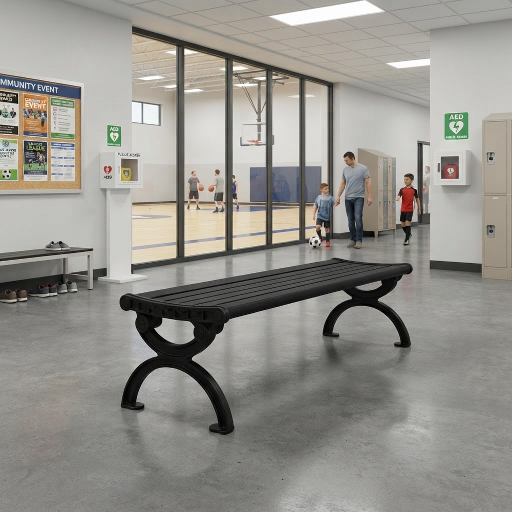A black outdoor-style bench with curved legs is placed in a modern indoor hallway. Large glass windows look into an indoor basketball court where people are playing.
