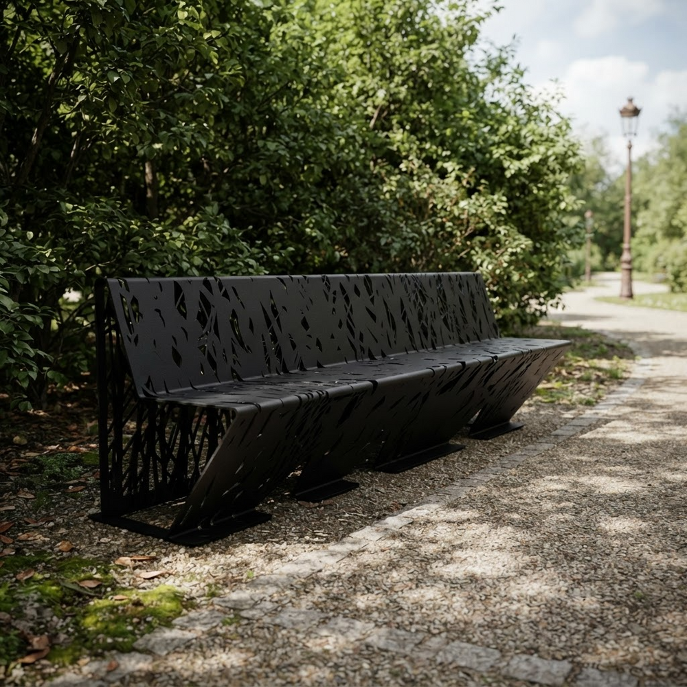 A modern black metal park bench with a leaf-cutout pattern, situated on a gravel path next to lush green trees.