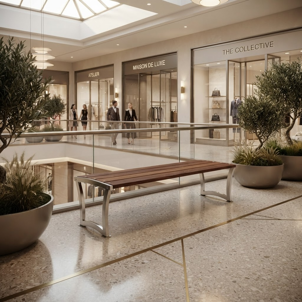 A modern wooden bench with sleek silver metal legs sits on a polished terrazzo floor in a luxury shopping mall atrium, flanked by potted olive trees and glass railings.