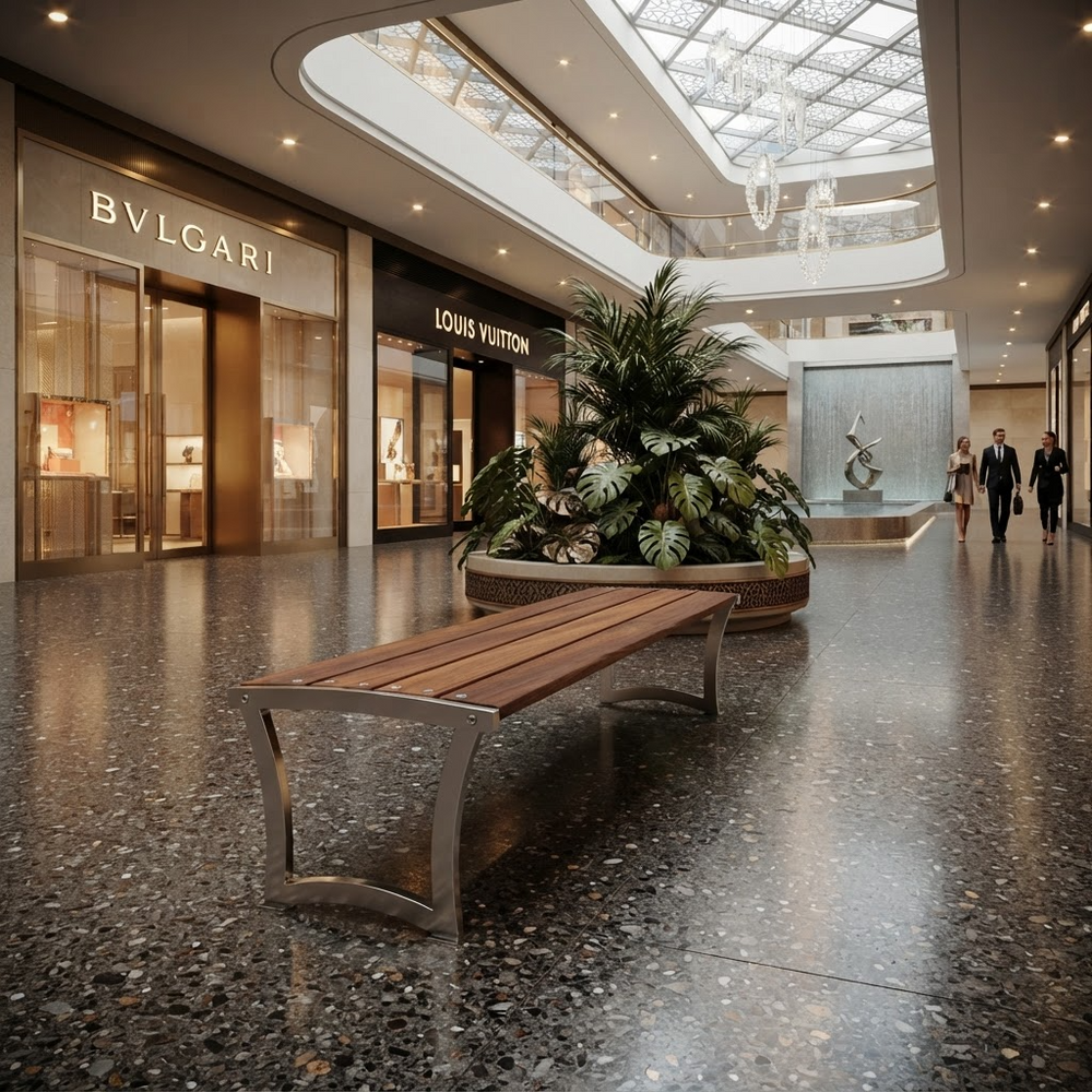 A modern wooden bench with a curved silver metal frame sits in a spacious, upscale shopping mall atrium with terrazzo floors, a large potted plant, and visible storefronts including Bvlgari and Louis Vuitton.
