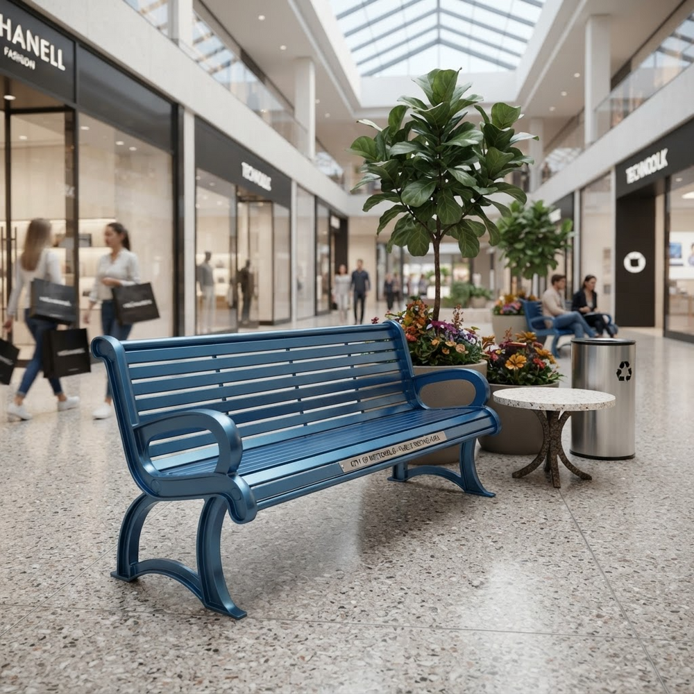 A blue slatted park bench in a modern shopping mall, shown with a small round table, a potted fiddle-leaf fig, and a stainless steel recycling bin. The mall interior features a high ceiling with skylights and various storefronts in the background.