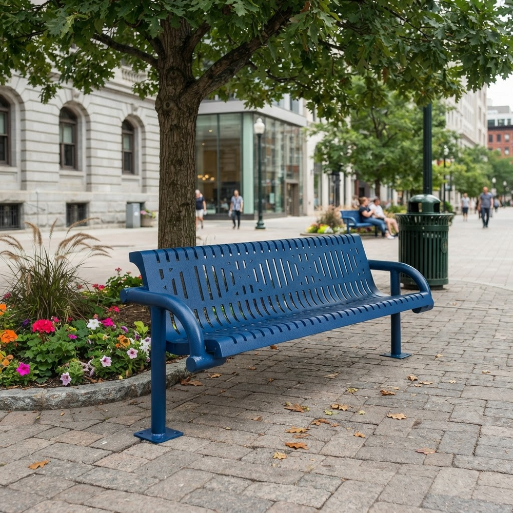 A blue metal park bench with a slatted back and seat, situated on a paved city sidewalk next to a tree with colorful flowers in a planter.