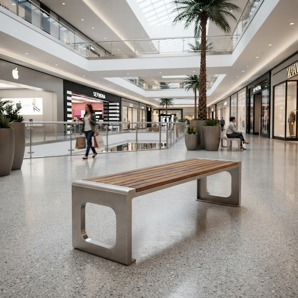 A modern wooden and metal bench in a spacious, sunlit shopping mall atrium with visible store fronts, palm trees, and walking shoppers in the background.