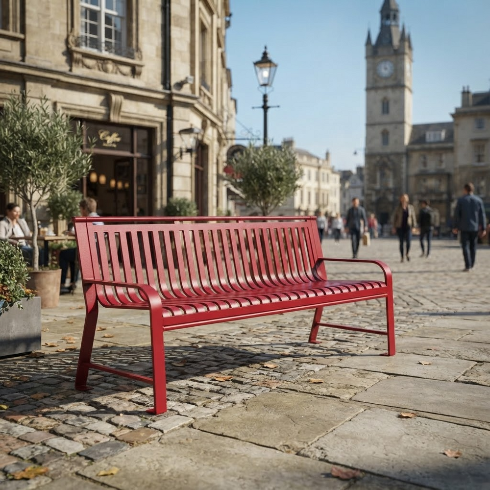 A bright red metal park bench with a slatted back and seat, positioned on a cobblestone street in a European town square with historic buildings and pedestrians in the background.