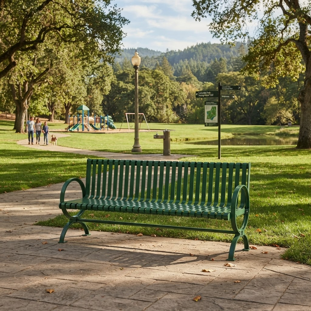 A green metal park bench sits on a stone pathway in a sunny, tree-filled park with a playground, people, and a pond in the background.