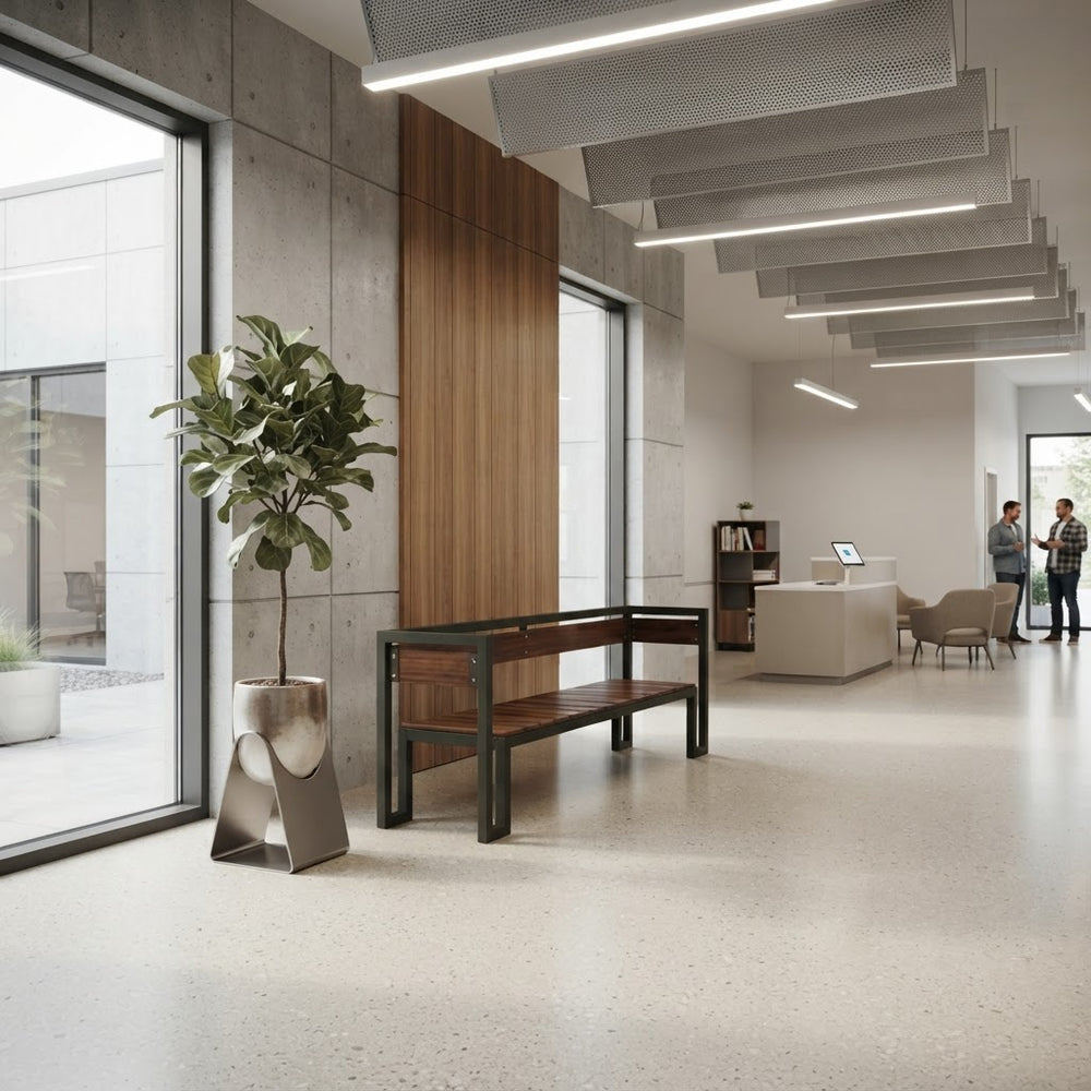 A modern office lobby with a wooden and metal bench in the foreground, a large potted plant, concrete walls, and a reception desk in the background. Two people are standing near the desk in the distance.