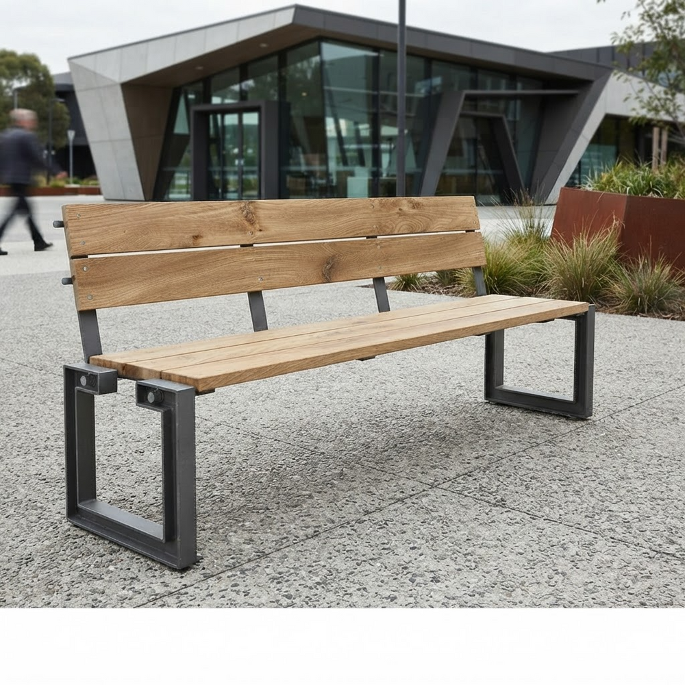 A modern outdoor bench with light natural wood slats and a black metal frame, situated on a paved plaza in front of a contemporary glass building.