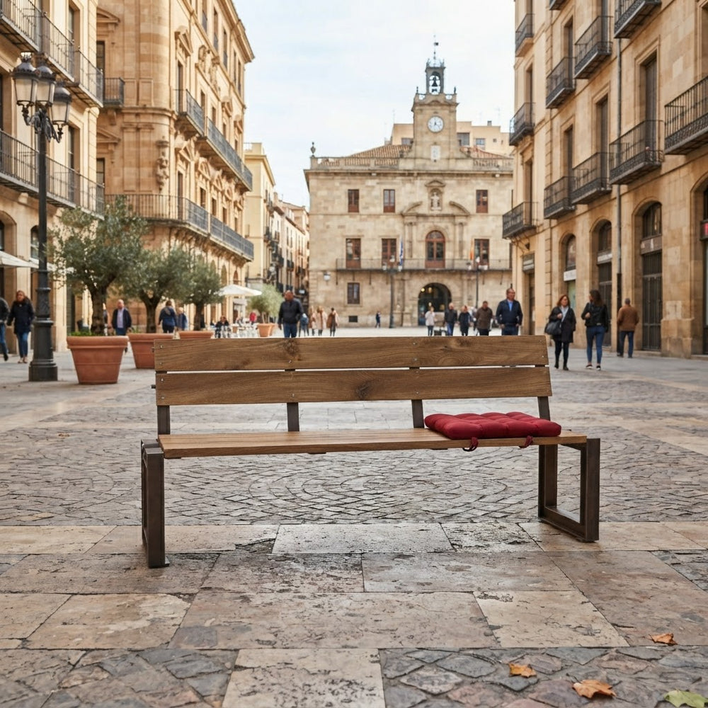 A wooden park bench with dark metal legs sits on a cobblestone plaza in a historic city square, with a red cushion resting on the right side. People walk in the background between tan buildings and a clock tower is visible in the distance.