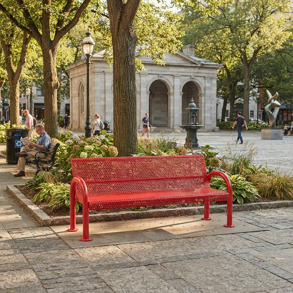 A bright red metal park bench with a perforated mesh seat and back, situated in a paved public square with trees, a stone building, and a few people in the background.
