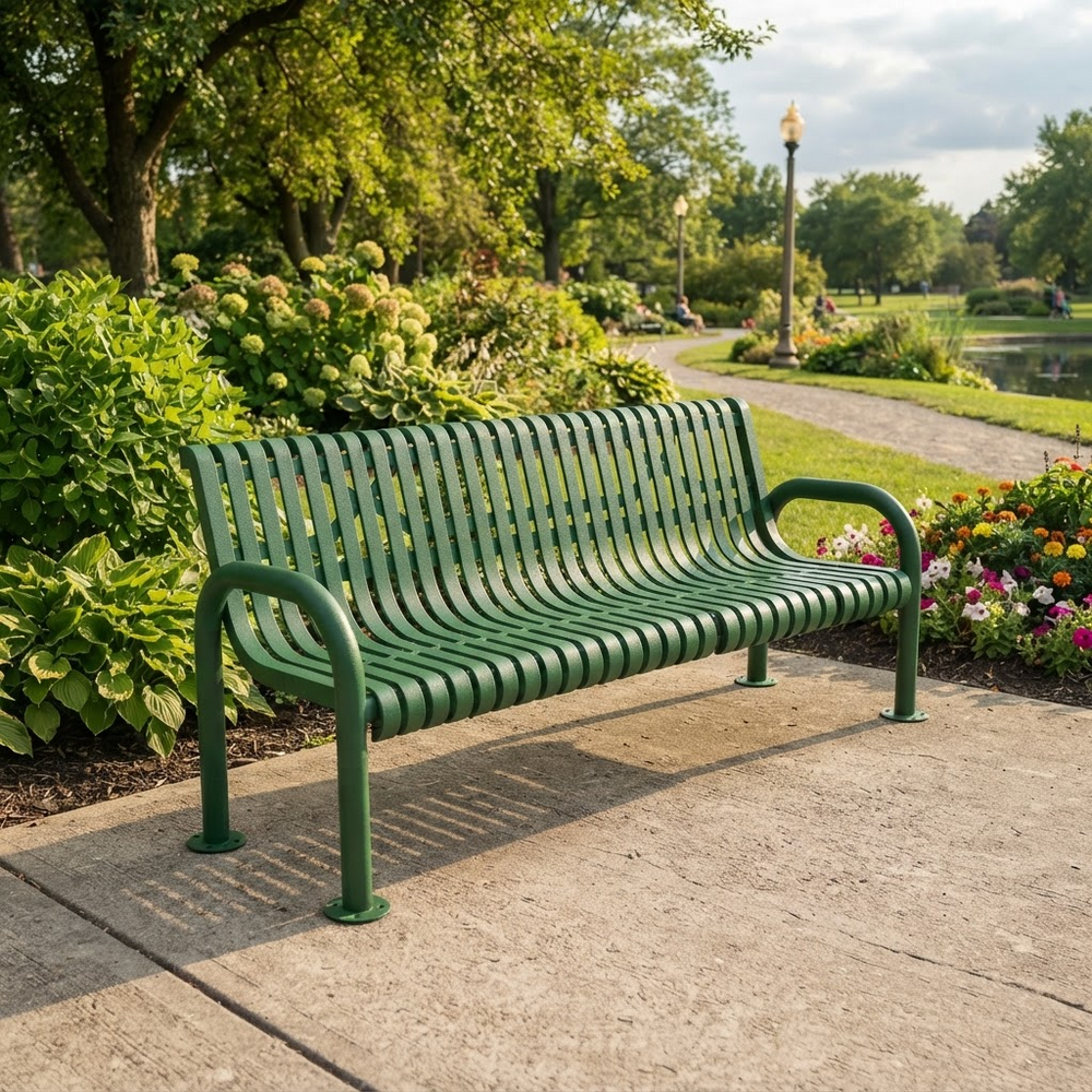 A green metal park bench with curved armrests sits on a concrete path in a landscaped garden with trees, flowers, and a pathway leading into the background.