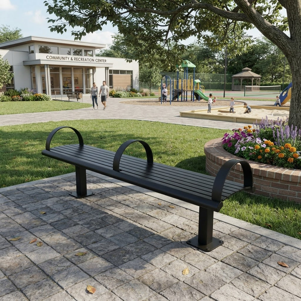 A black metal park bench with curved backrests sits on a stone patio in a landscaped outdoor area. In the background, there is a grassy lawn, a children's playground, a flower bed, and a building labeled 'Community & Recreation Center'.