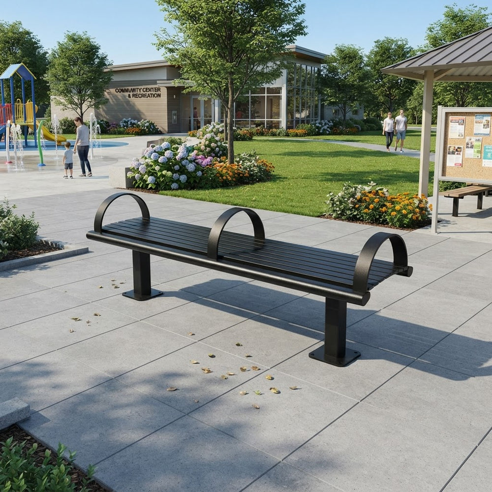 A modern black metal park bench with two curved backrests, situated on a paved outdoor plaza in a landscaped community center area with grass, trees, flowers, and nearby structures.