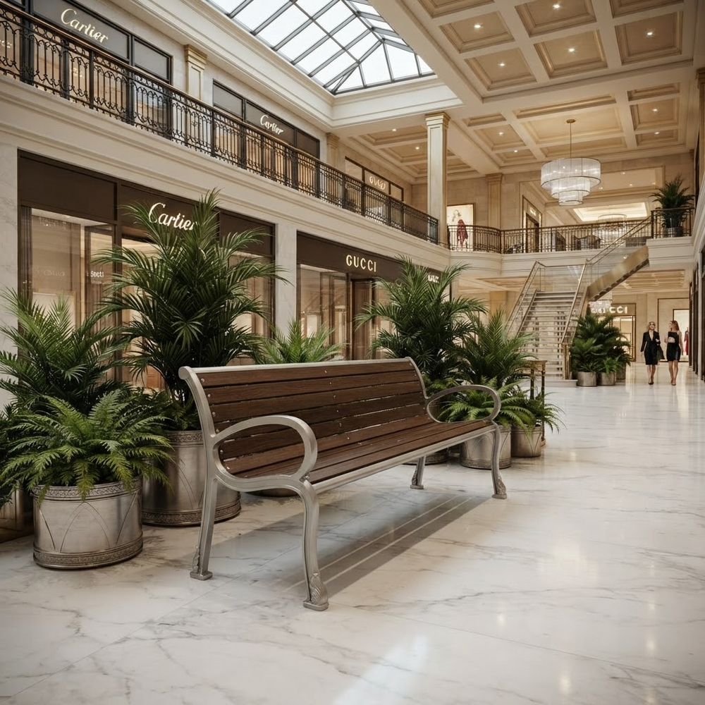 A wooden slat bench with a silver metal frame sits in a grand, upscale shopping mall atrium with marble floors, potted plants, and visible Cartier and Gucci storefronts in the background.