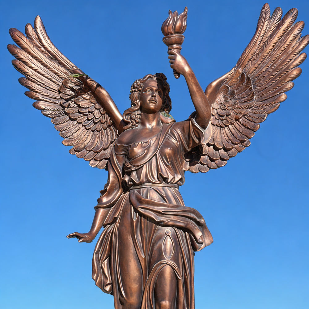 Bronze statue of an angel holding a torch against a clear blue sky
Bronze angelic figure with wide wings and torch
