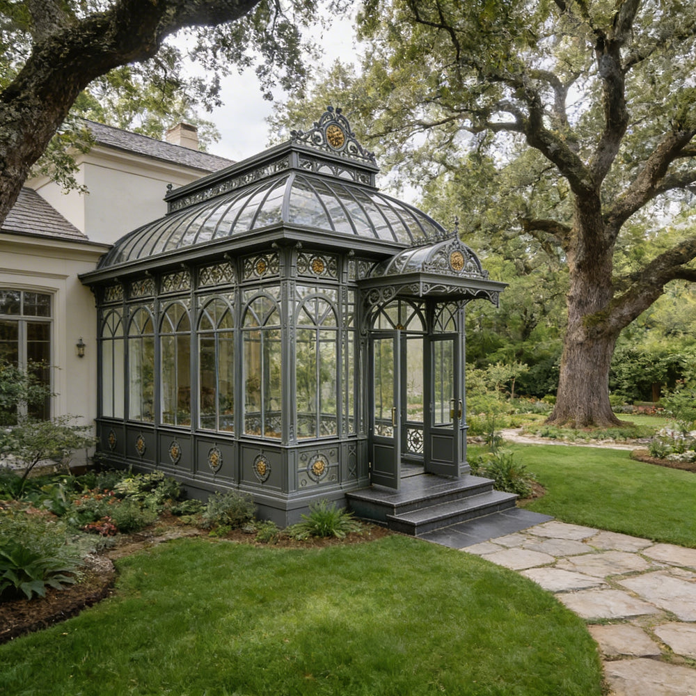 Victorian-style glasshouse in a garden setting with trees and a house in the background.
