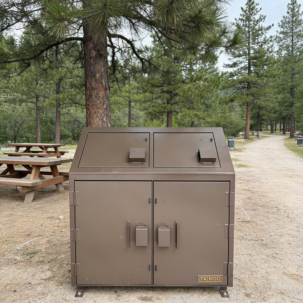 Brown metal trash bin with 'Taimco' branding in a park setting with trees and picnic tables.