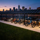 Row of electric bikes parked under a shelter with a city skyline in the background at dusk.