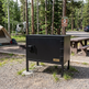 Black metal camp oven with 'Taimco' branding in a camping setting with trees and picnic table.