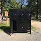 Black metal lockers with one door open, set against a natural background with trees and a path.