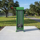 A green metal bicycle pump and tire repair kit stands on a concrete pad in an outdoor park setting with trees, grass, and a paved path in the background.