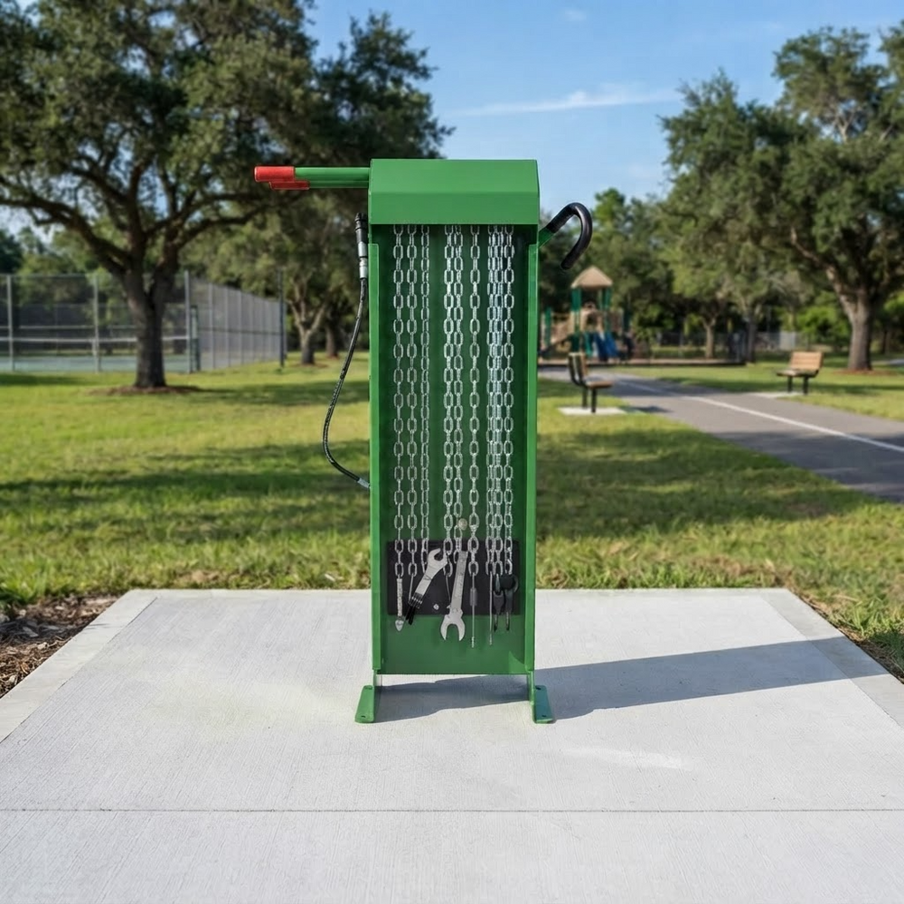 A green metal bicycle pump and tire repair kit stands on a concrete pad in an outdoor park setting with trees, grass, and a paved path in the background.