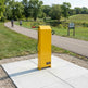 A bright yellow park ticketing machine mounted on a square concrete pad beside a paved path in a park, with trees, grass, and a small pond in the background. A small sign on the right reads Park Ticket.
