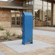 A blue metal public library card reader kiosk stands on a light concrete pad outside a brick and glass City Public Library building. The device has a red handle, a black cable, and a small gold-toned brand plaque near the bottom.