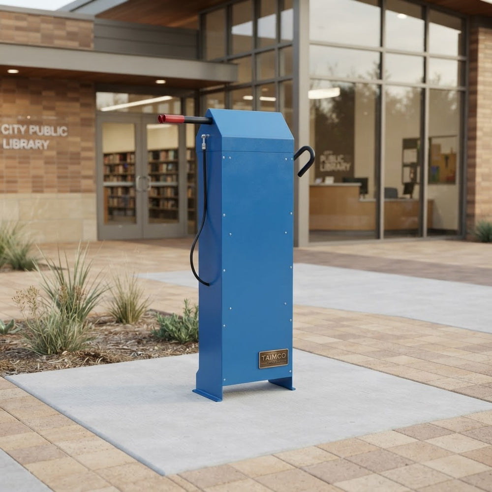 A blue metal public library card reader kiosk stands on a light concrete pad outside a brick and glass City Public Library building. The device has a red handle, a black cable, and a small gold-toned brand plaque near the bottom.