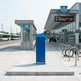 A blue public transit payment kiosk or fuel-style machine stands on a concrete platform outside a modern bus station. The station building has a glass facade, a digital departure board, bike racks with bicycles, and another shelter structure in the background.