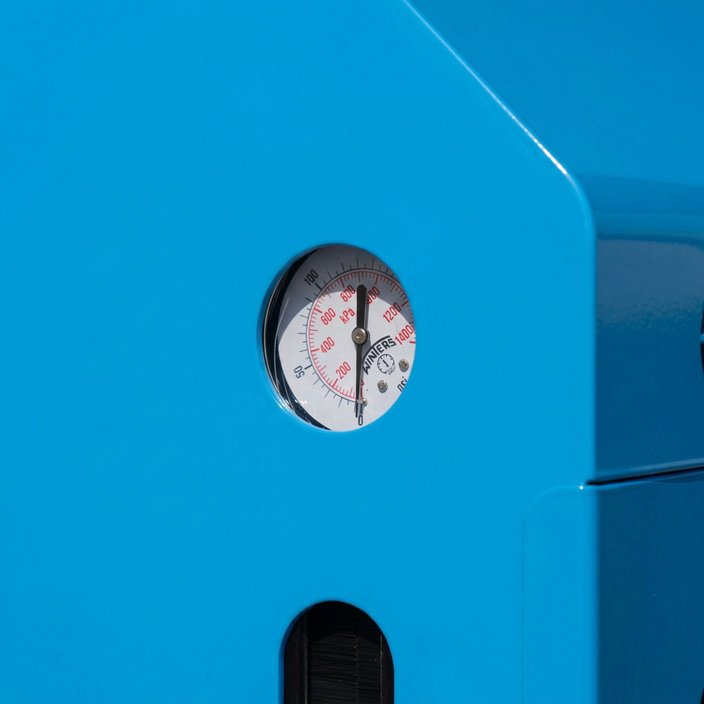 Close-up of a bright blue appliance or machine with a circular pressure gauge visible through a cutout. The gauge face is white with red markings and a black dial.